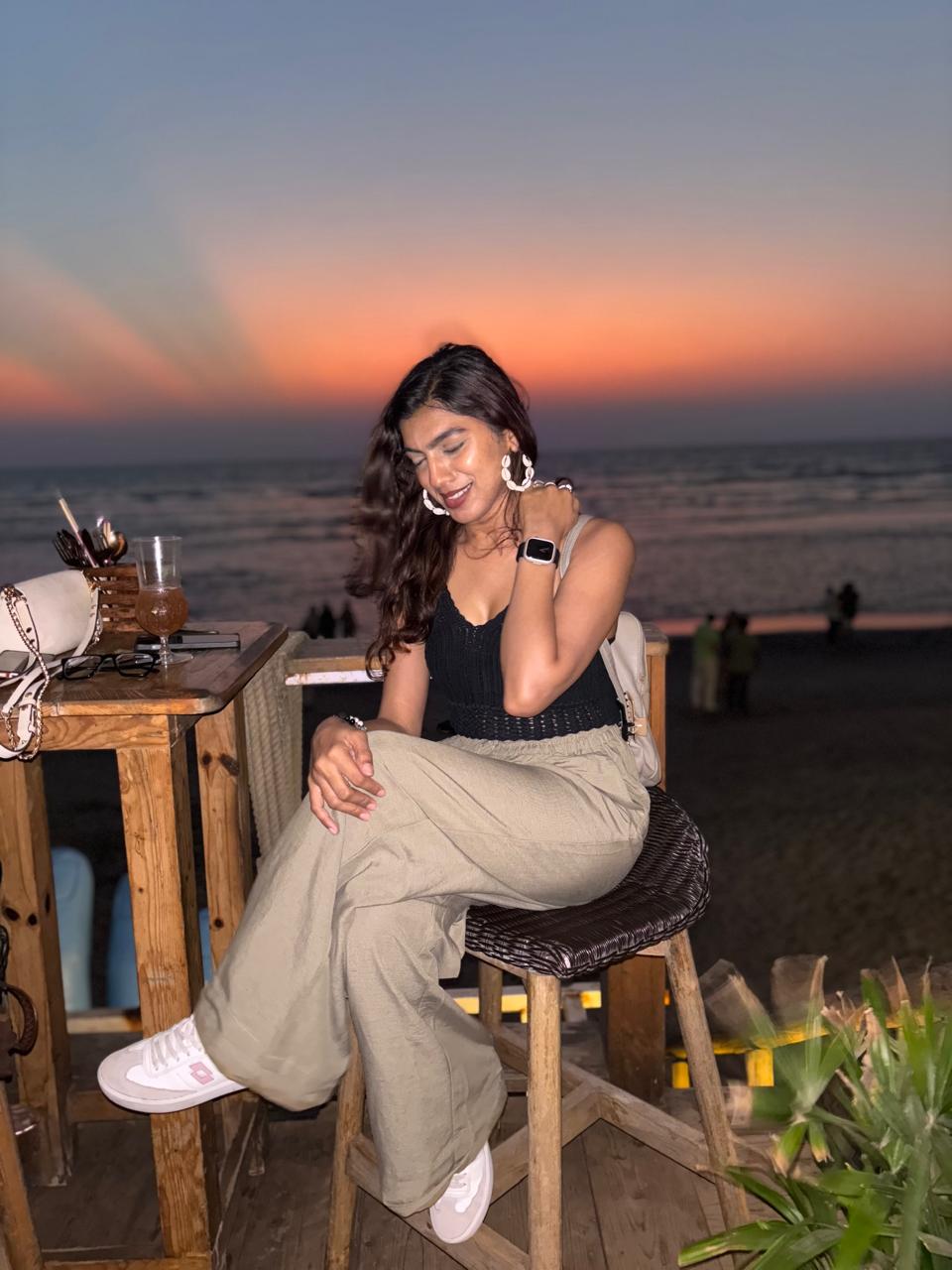Woman sitting on a stool at sunset, smiling, wearing a black top and beige pants, with a beach in the background.