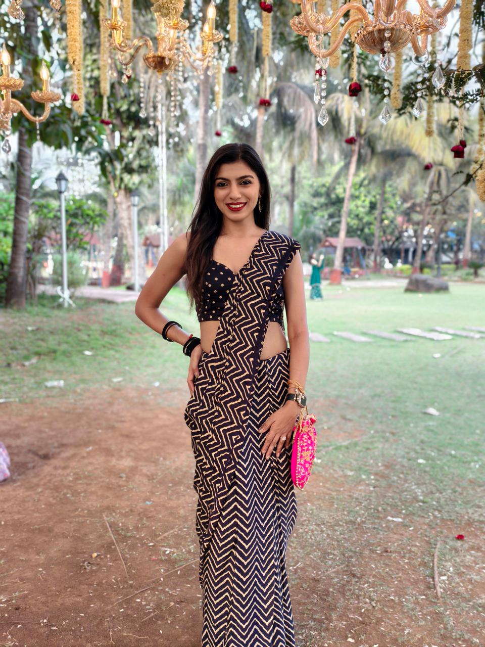 Woman in patterned dress holding pink clutch, standing outdoors with hanging lights and greenery in background.