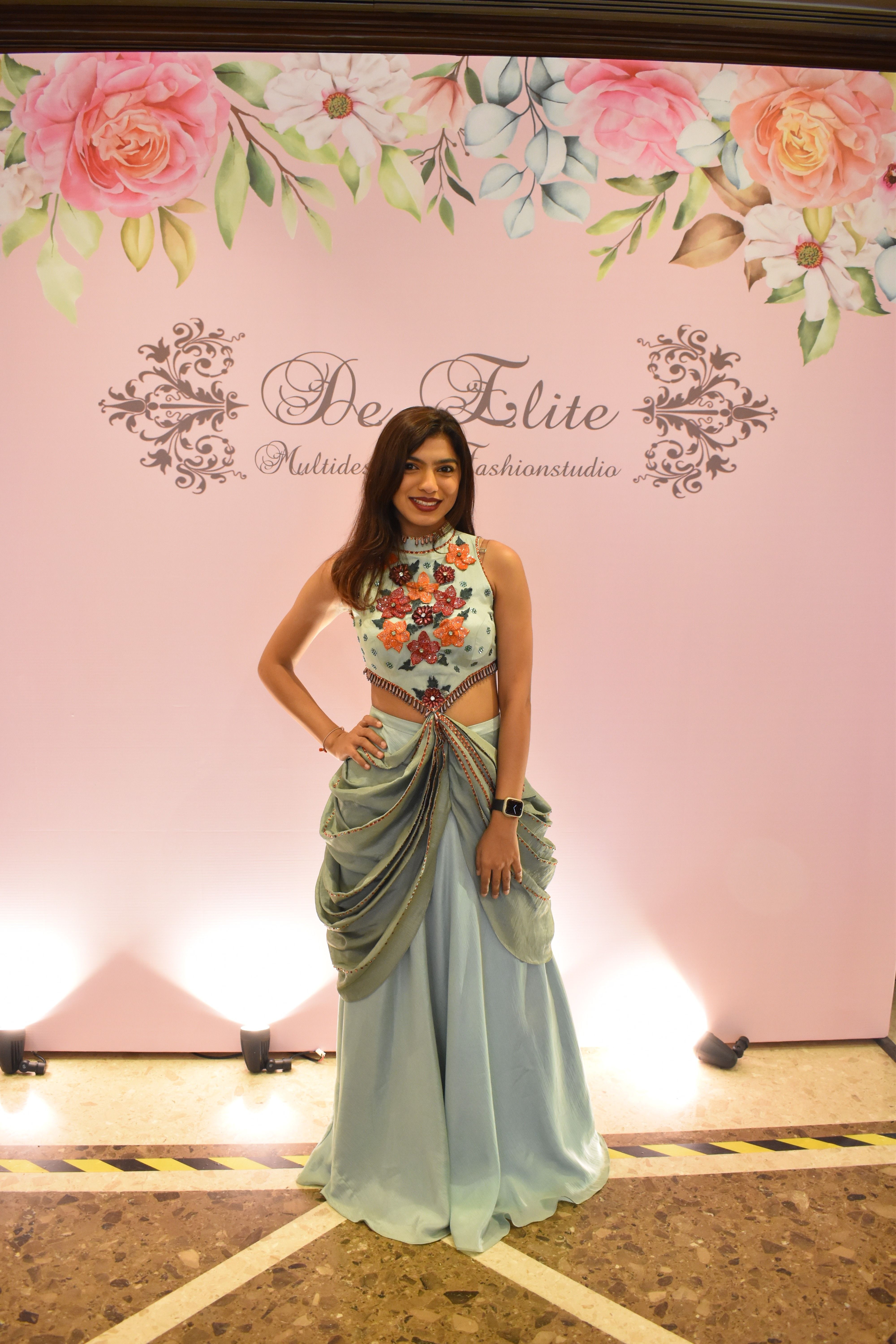 Woman in a floral top and long skirt standing on a stage with pink floral backdrop, smiling.