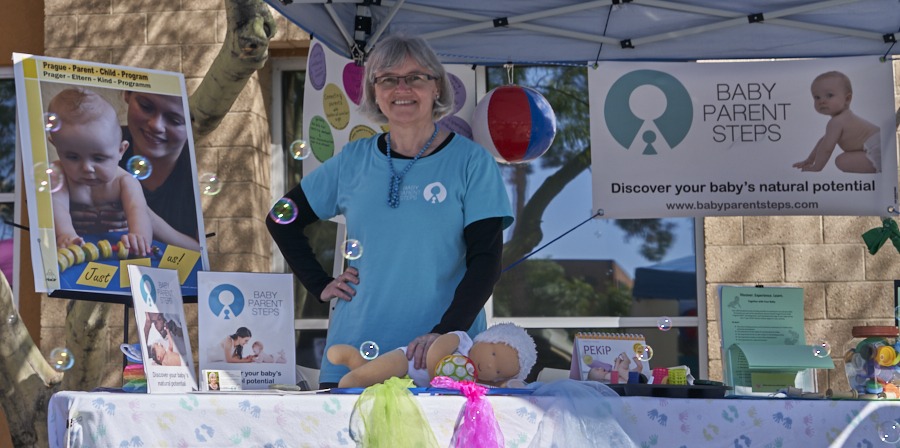 Woman standing at an outdoor booth with baby-related informational materials and a banner, smiling, with trees in background.