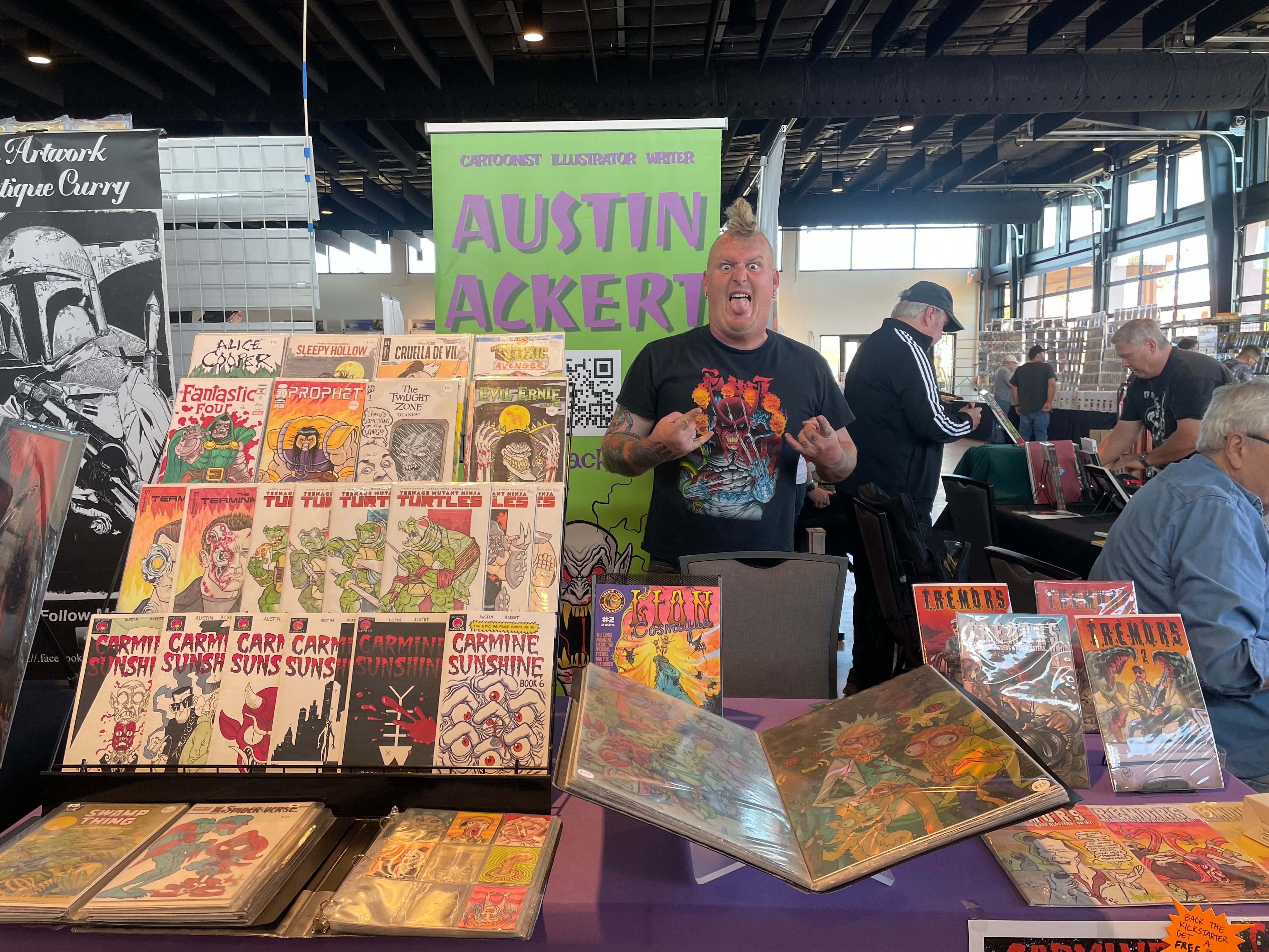 Person standing behind a table with comic books and graphic novels at a convention or event, with a green sign in the background.