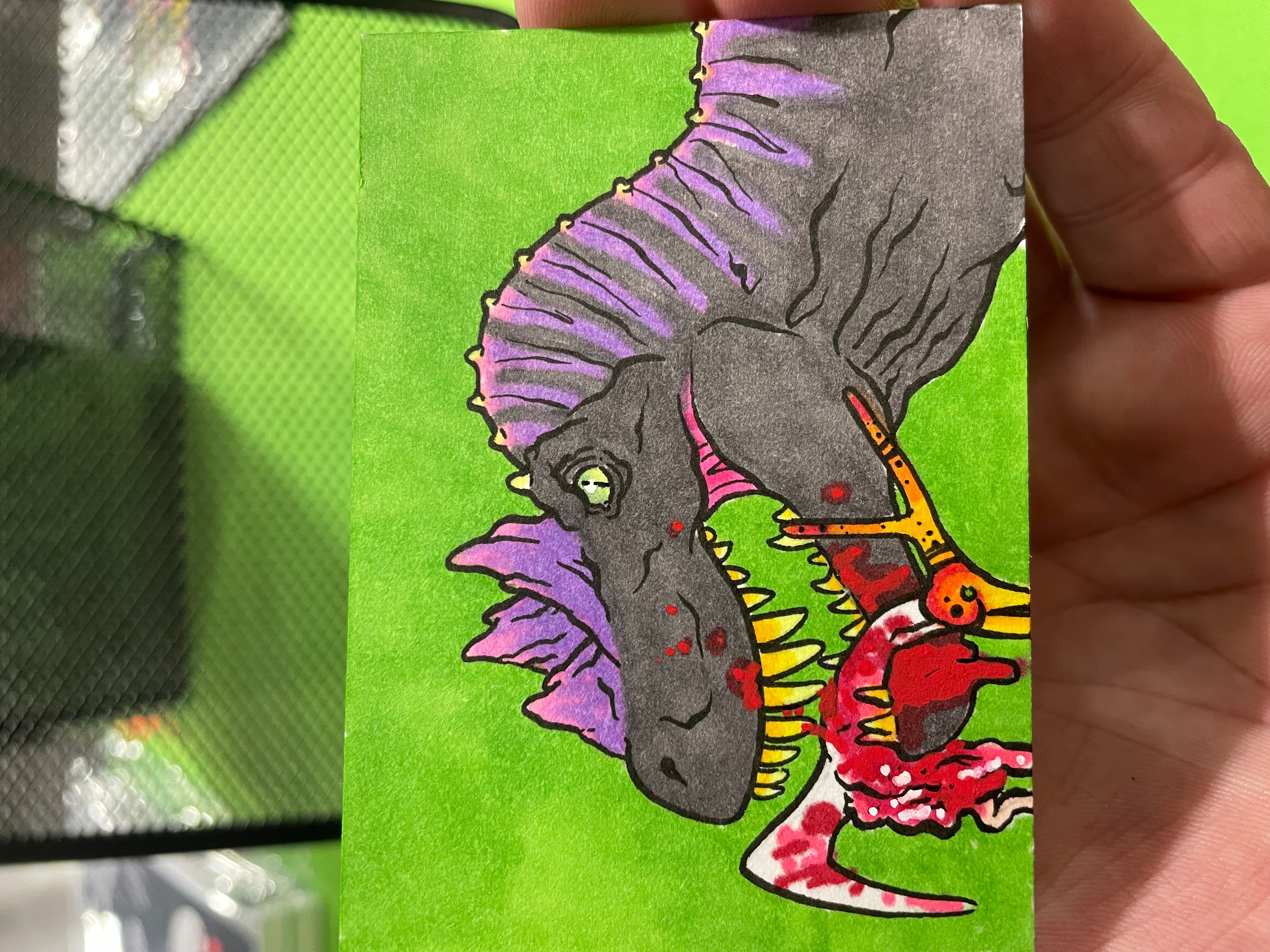 Hand holding a colorful drawing of a dinosaur and a bird on a green background.