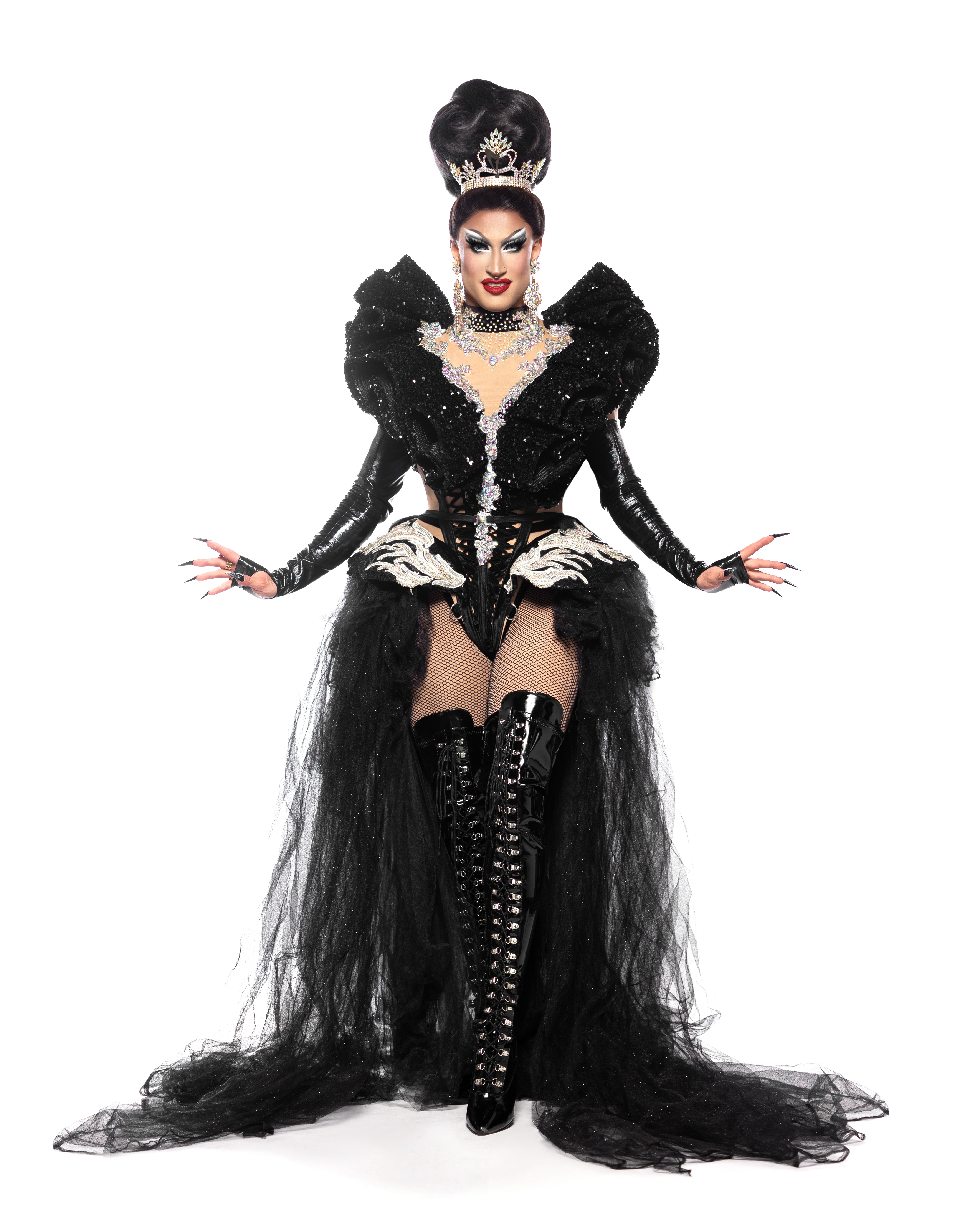 Woman in elaborate black gown with high hair, lace-up boots, and dramatic makeup, posing confidently against a white background.