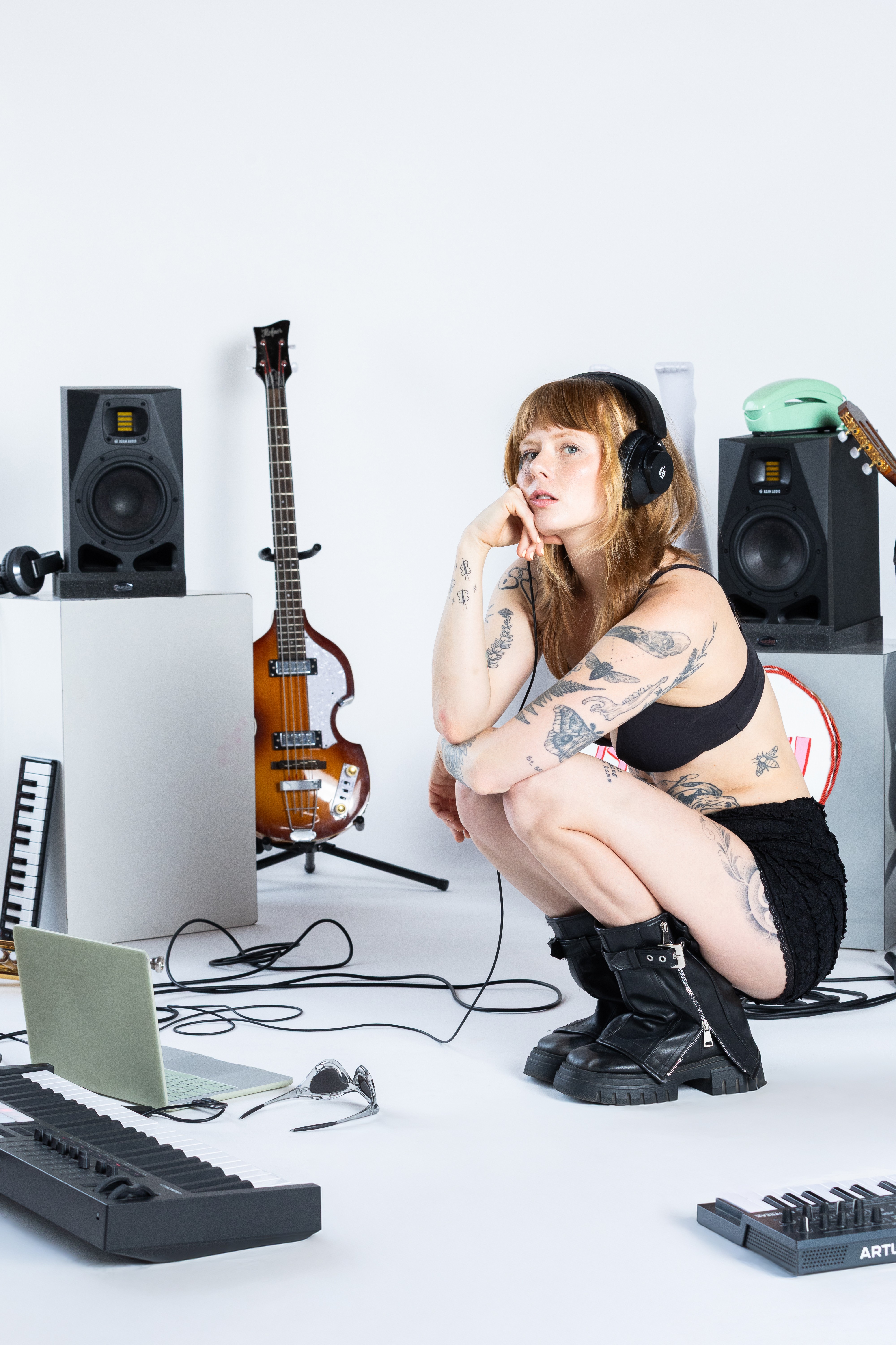 Young woman with headphones crouching among music equipment, including speakers, guitars, and keyboards, in a bright studio.