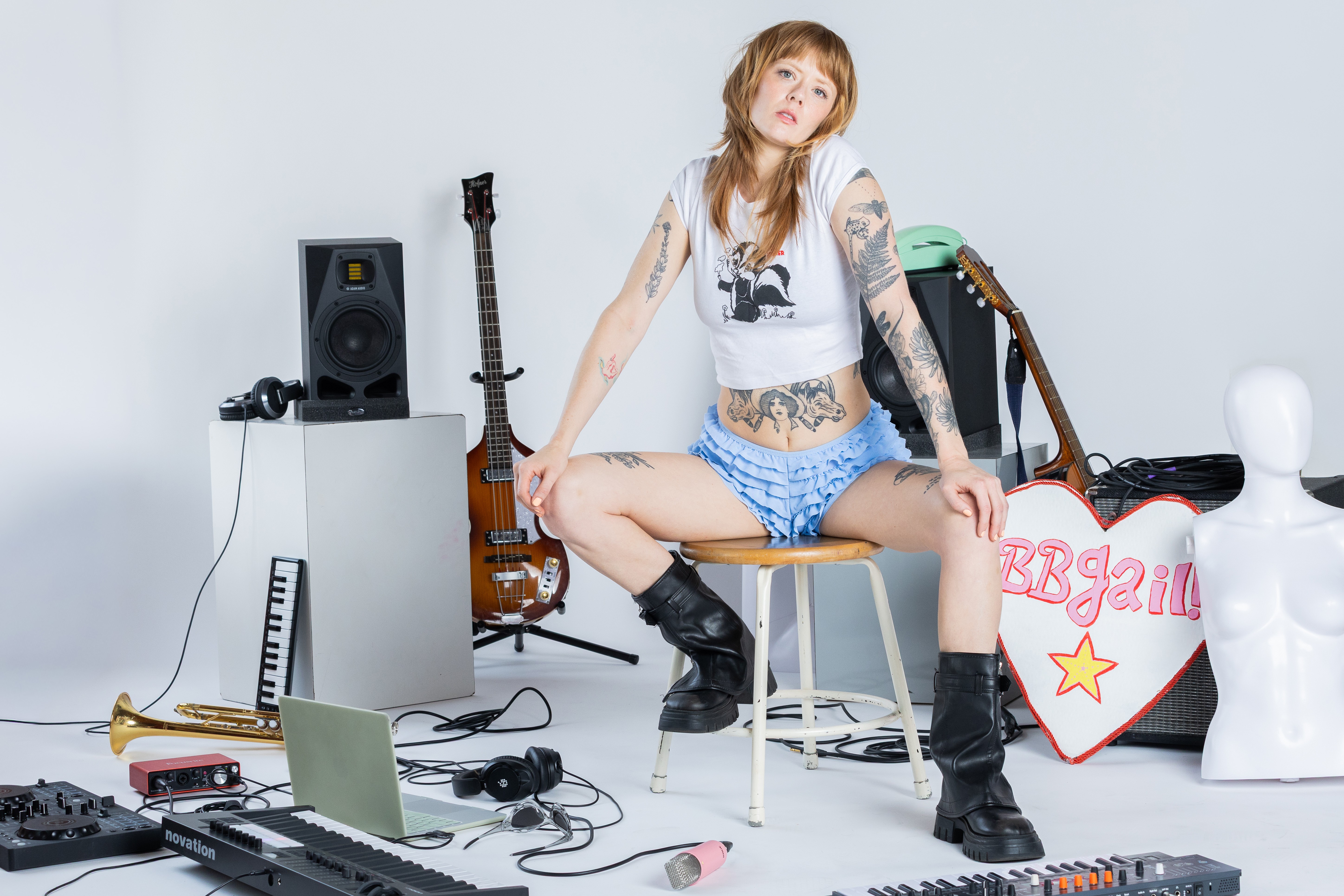 Young woman with tattoos sitting on a chair surrounded by musical instruments and equipment, with a white mannequin and a bag in the background.