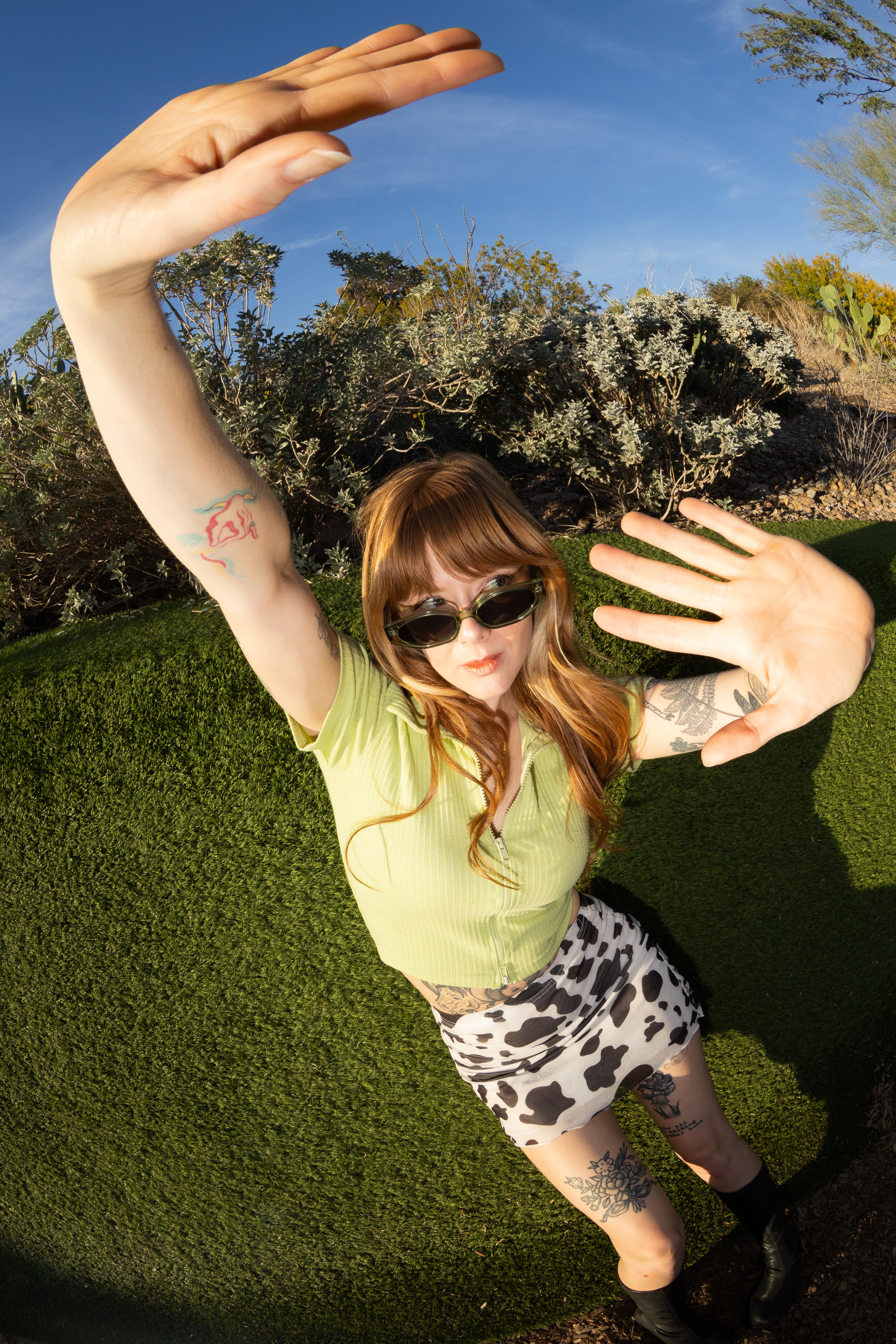 Person with sunglasses and patterned shorts raising arms outdoors, with trees and blue sky in background.
