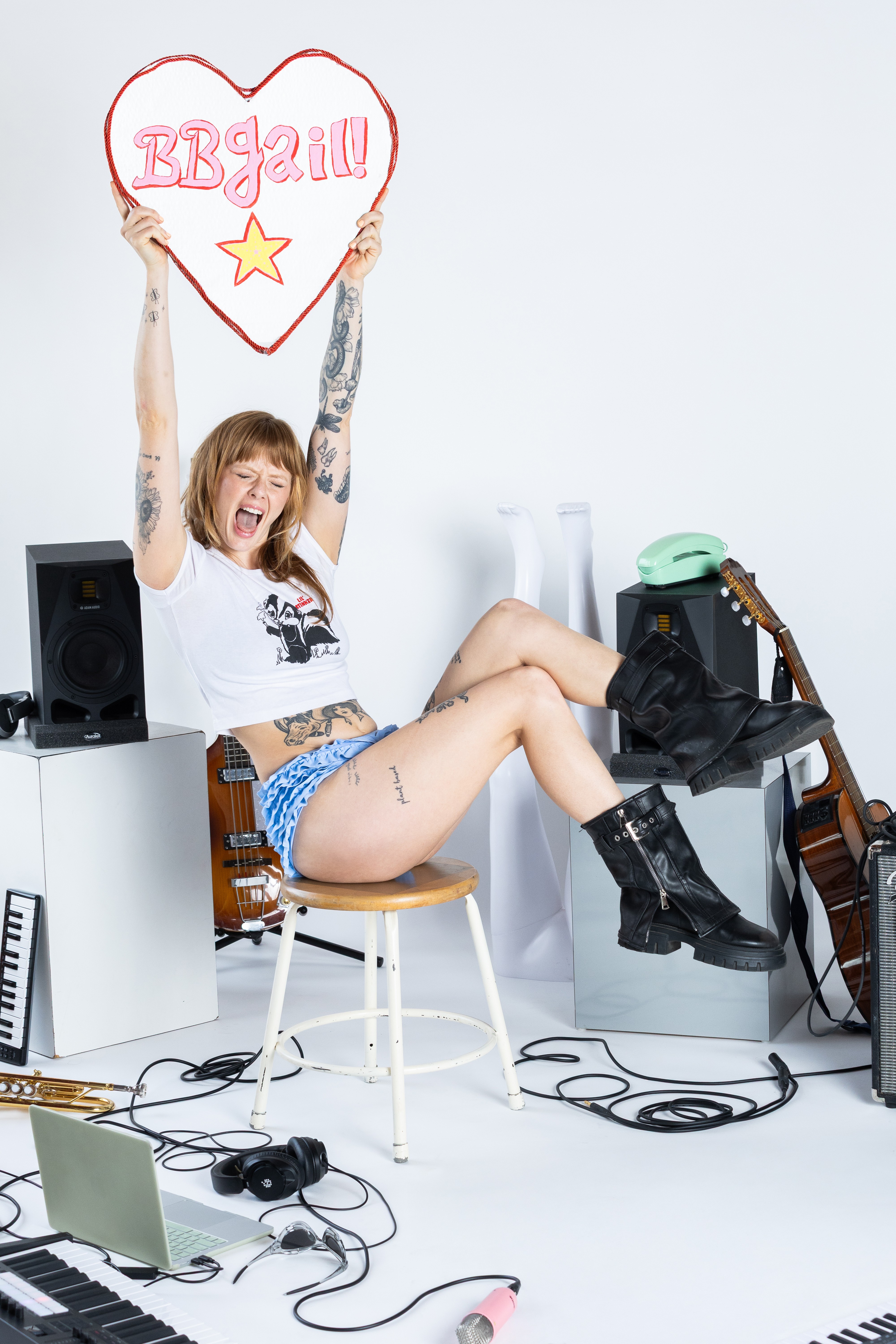 Young woman with tattoos holding a heart-shaped sign with a star, sitting on a chair surrounded by musical equipment, smiling.