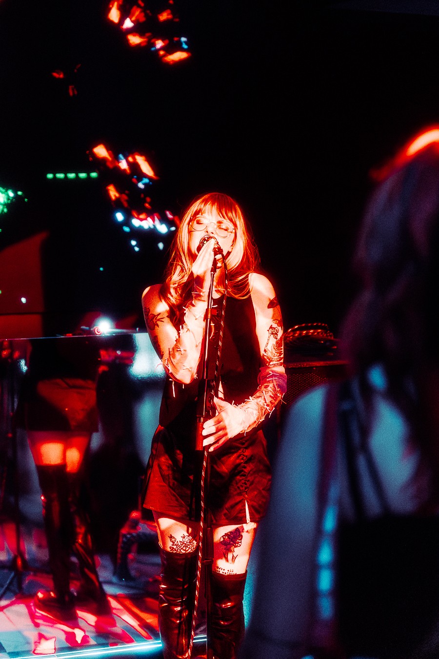 A woman with long hair and glasses singing into a microphone on stage with colorful lights, in a dark setting.