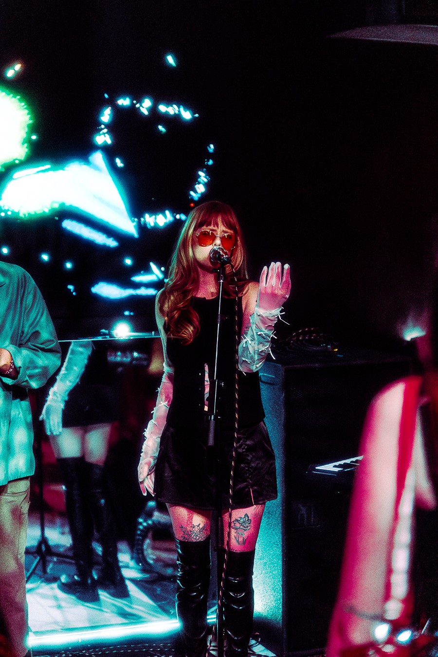 A woman with long hair and sunglasses performs on stage with a microphone, surrounded by colorful lights and other performers.