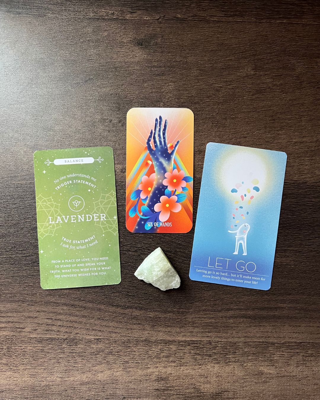 Three cards and a white stone on a wooden surface, with the middle card showing a hand with flowers and rainbow colors.
