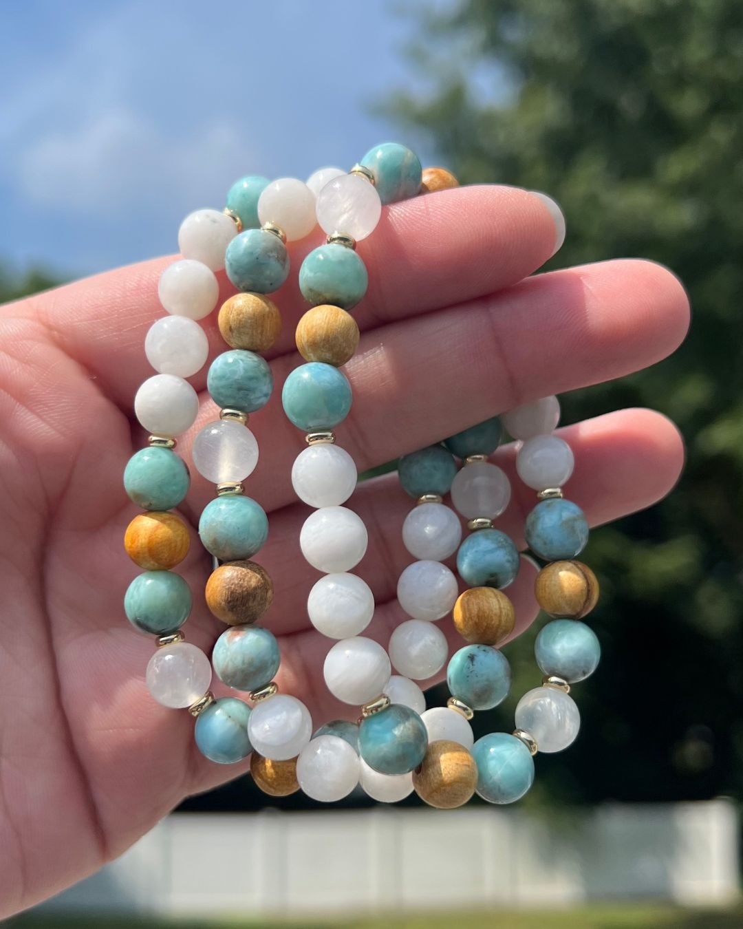 Hand holding three beaded necklaces with white, turquoise, gold, and wood-colored beads, outdoors with trees and sky in background.
