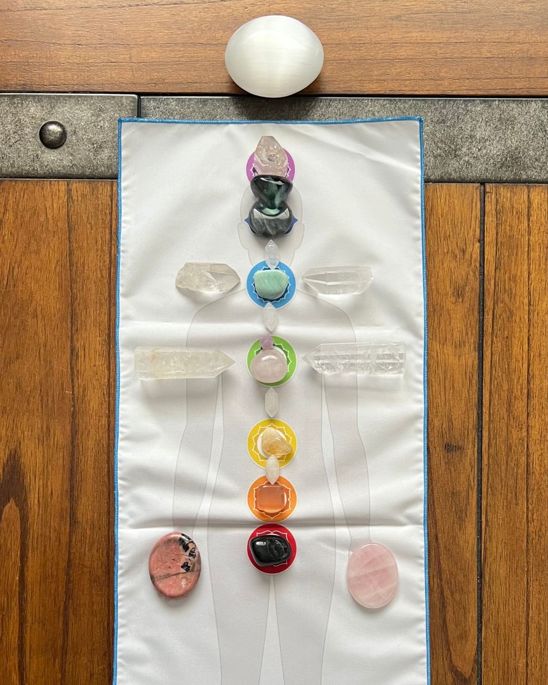 Egg on top, colorful pacifiers and pacifier clips arranged vertically on a cloth on wooden floor, with two pink objects at bottom.