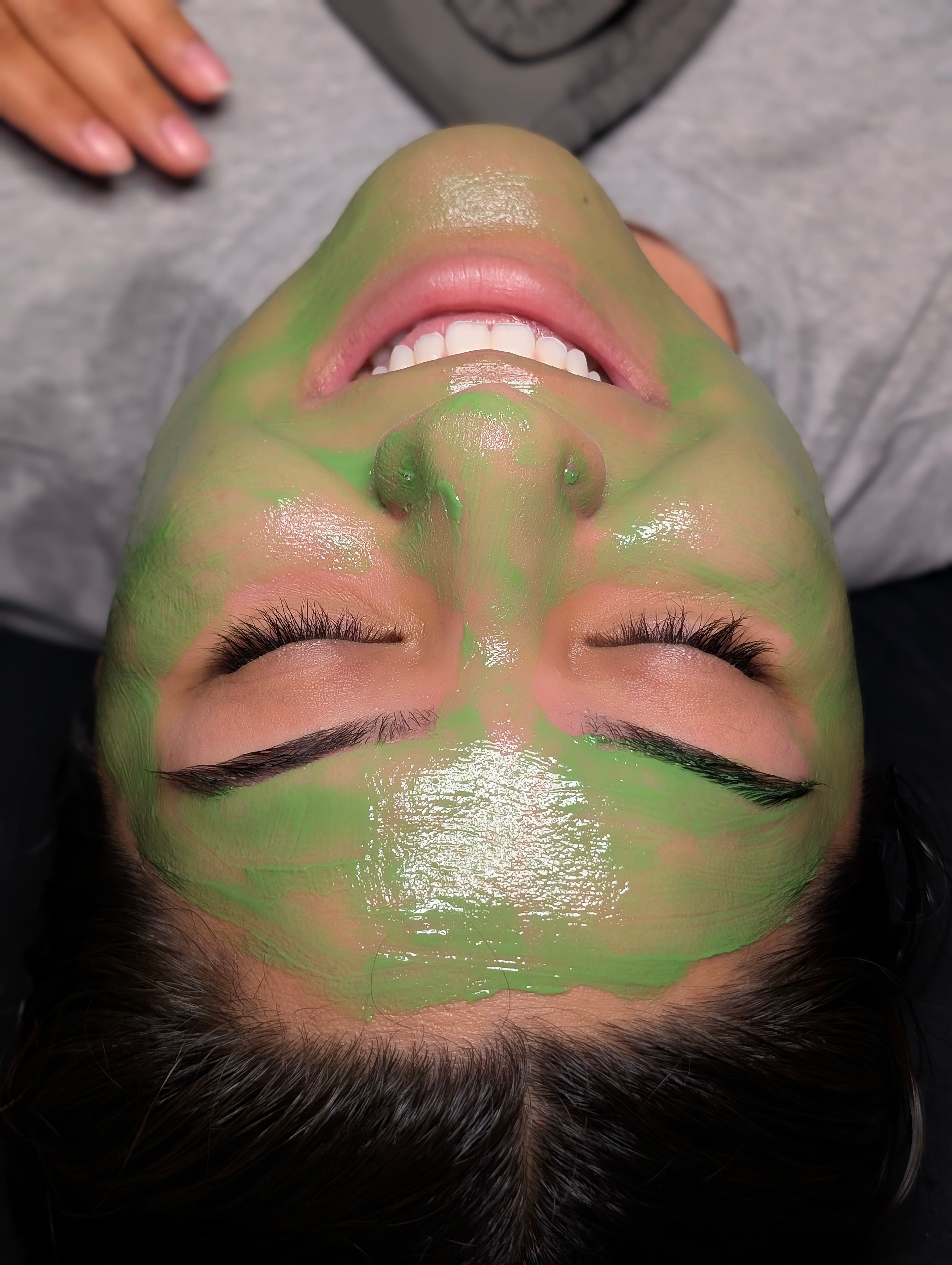 Person lying down with green facial mask, eyes closed, smiling, head tilted back, hair spread out.