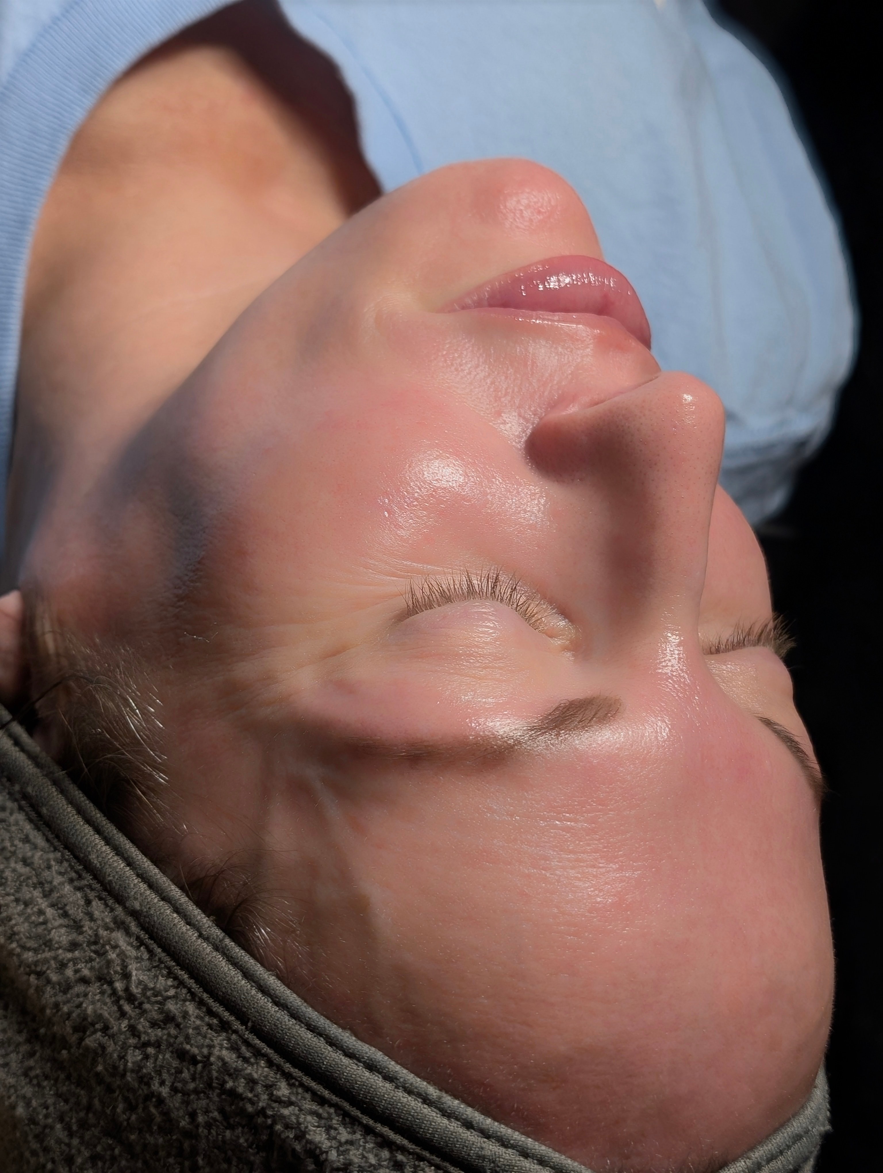 Person lying down with eyes closed, face visible, wearing a blue garment, head resting on a surface.
