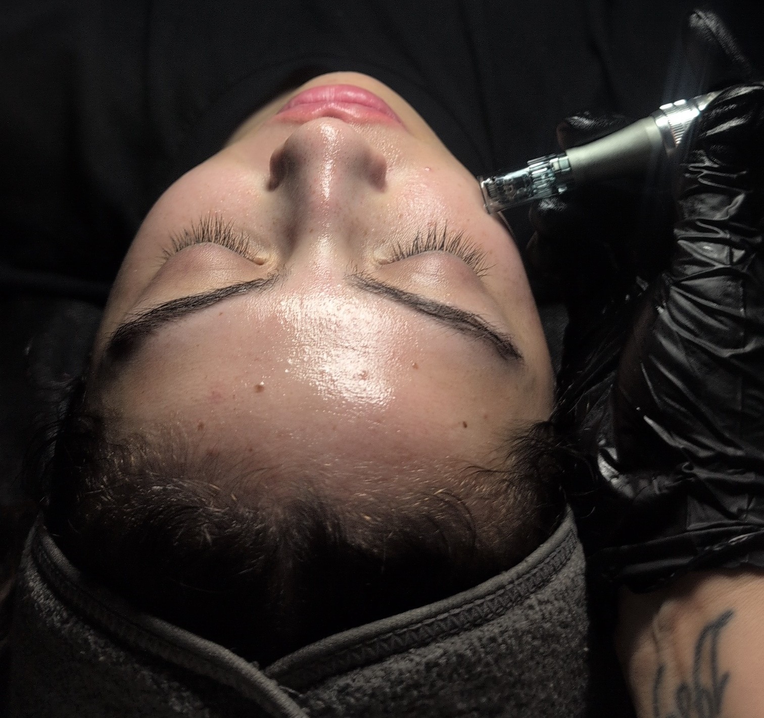 Person receiving a facial treatment with eyes closed, lying down, with a gloved hand holding a device near face.