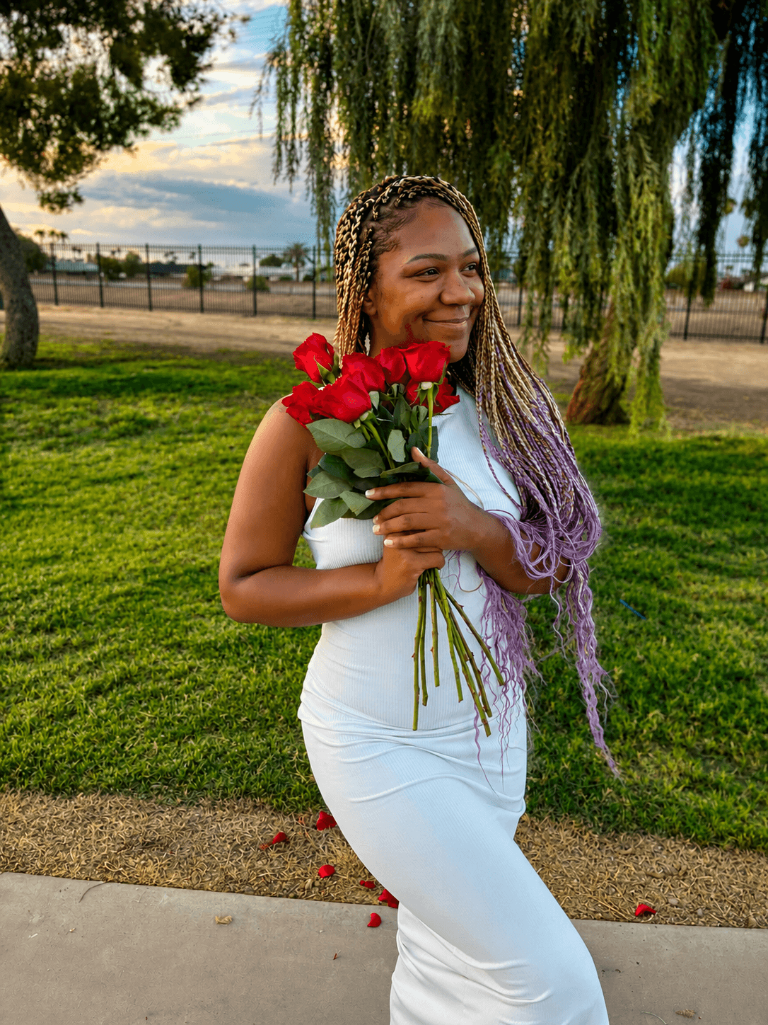 Woman in white dress holding a bouquet of red roses outdoors, smiling, with trees and sky in background.