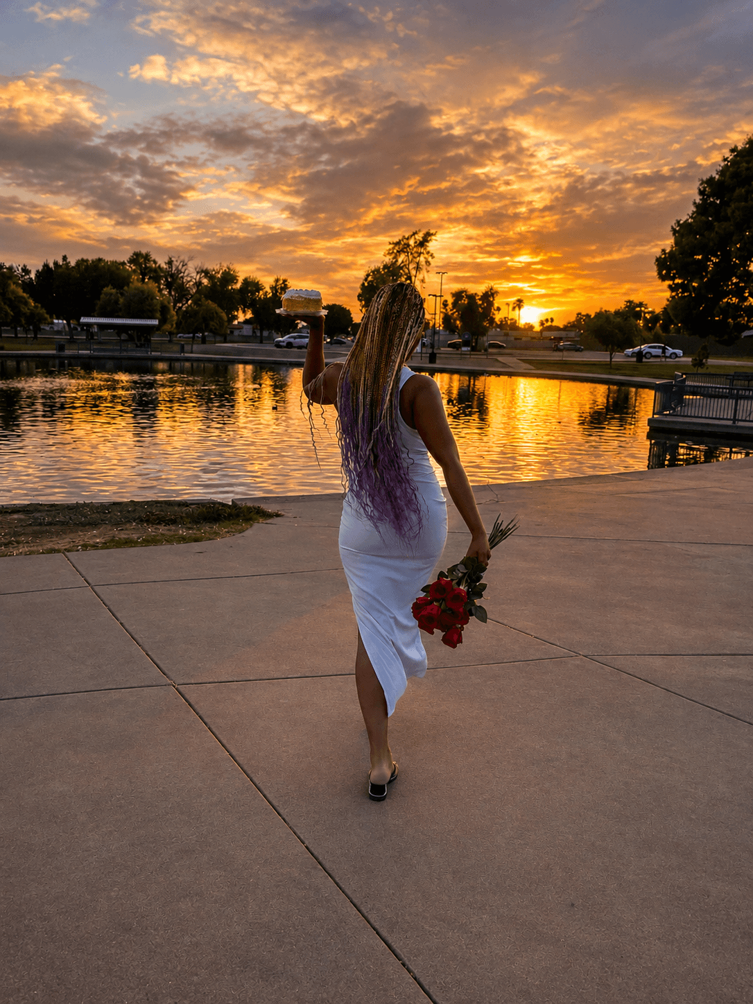 Woman with long hair holding a bouquet of red flowers walking near a lake at sunset, trees and sky in background.