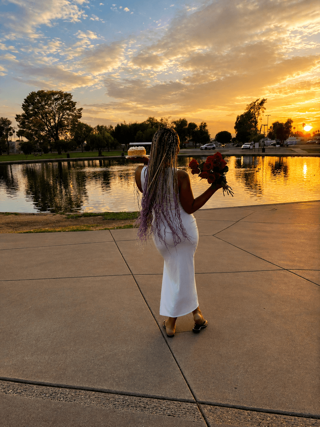 Woman with long hair in a white dress holding a bouquet by a lake during sunset, with trees and sky reflected in water.