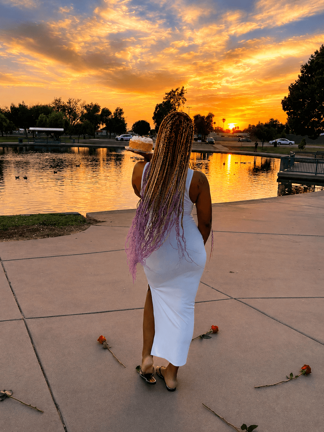 Woman with long hair in a white dress walking near a lake at sunset, with roses on the ground.